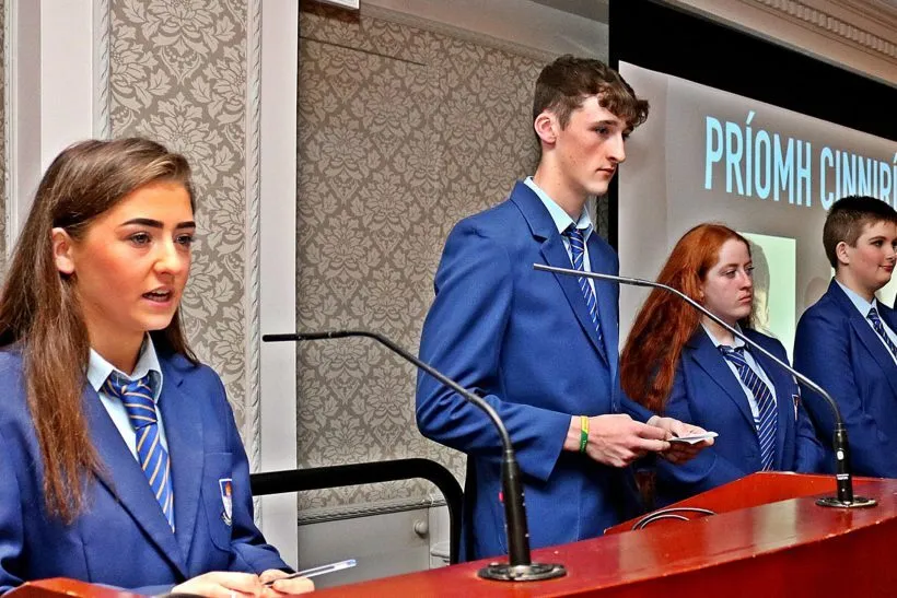Un grupo de estudiantes del St. Mogues College está de pie frente a un fondo con la inscripción Priory Church.