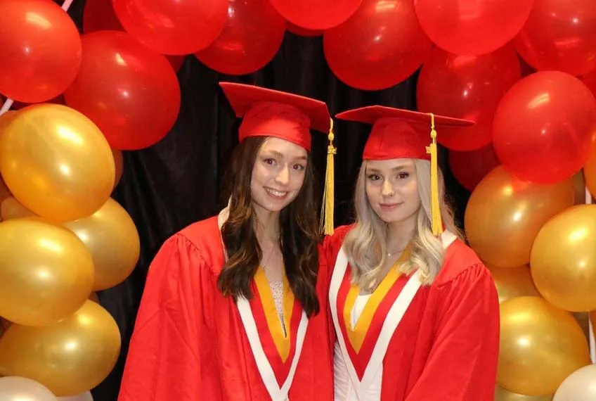Dos graduadas de Hammarskjold High School posan con sus vestidos de graduación frente a globos rojos y dorados.