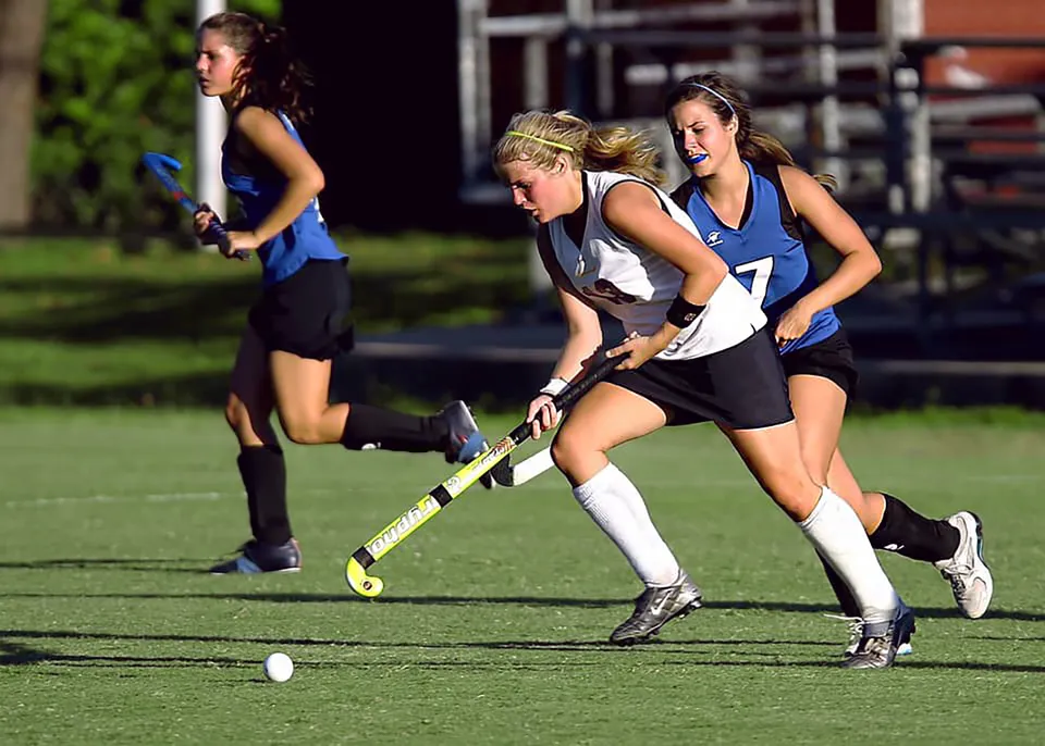 Studentesse della Frances Kelsey Secondary School giocano a hockey su prato su un campo sportivo con lo stadio sullo sfondo.