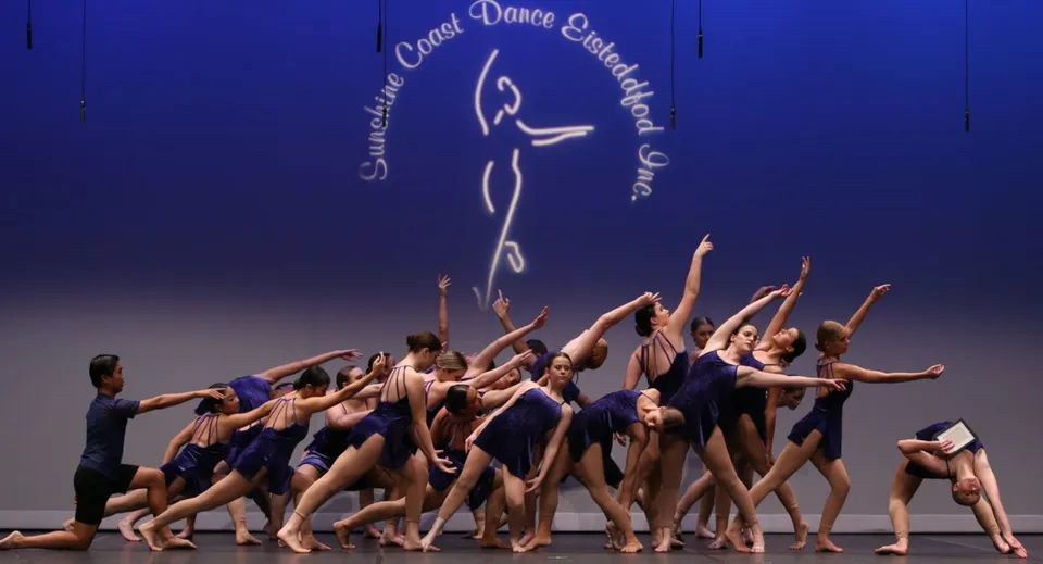 Un gruppo di ballerini si esibisce sul palco davanti a un grande logo della Sunshine Coast Dance Company alla Caloundra State High School.