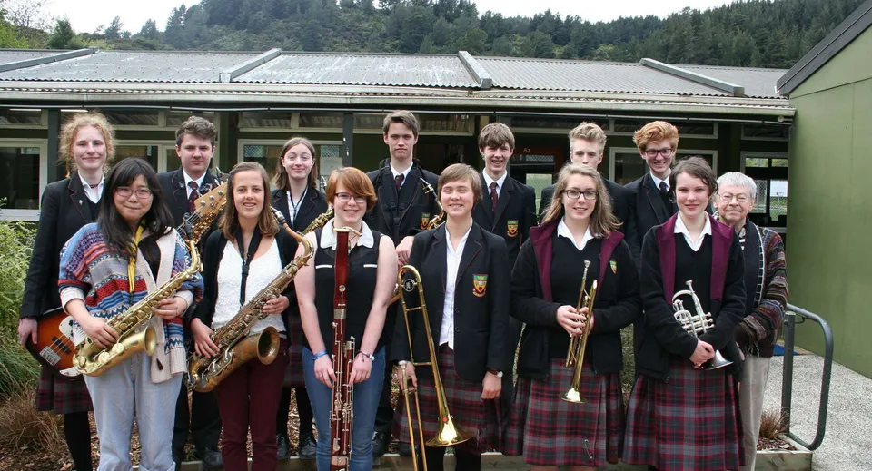 Un gruppo di studenti della Logan Park High School con strumenti musicali si trova davanti a un edificio, con uno sfondo boscoso.