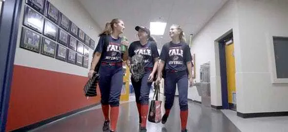 Tres estudiantes con uniformes de sóftbol azul y rojo caminan sonriendo por el pasillo