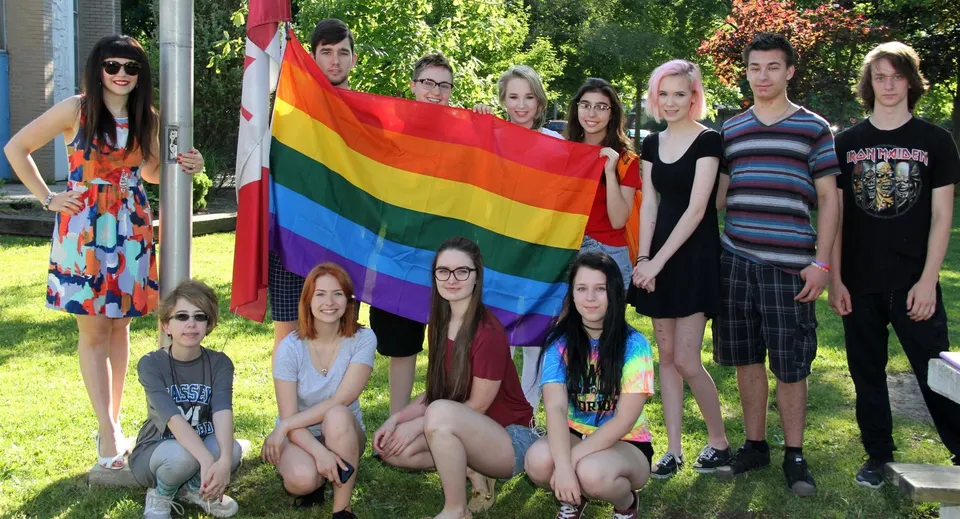 Un gruppo di studenti della Sandwich Secondary School posa all'aperto con una bandiera arcobaleno in un ambiente simile a un parco.