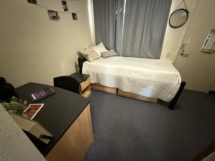 Habitación de la residencia con cama individual, escritorio oscuro y espejo redondo en la pared