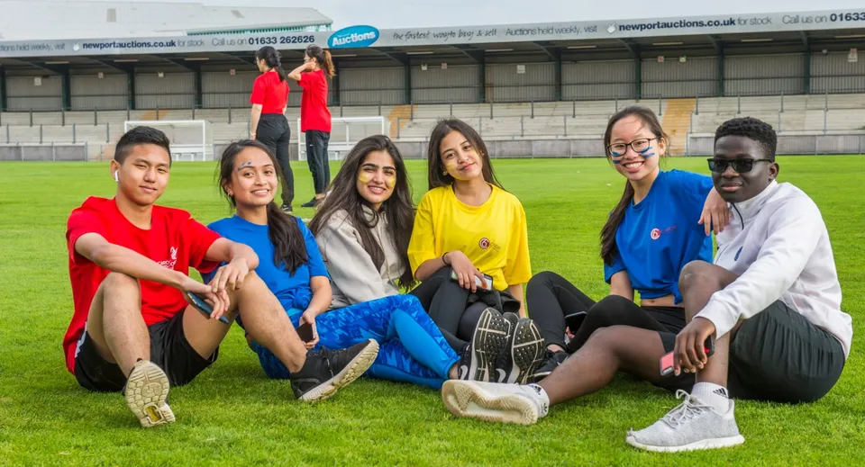 Un gruppo di studenti e studentesse siede su un prato davanti a uno stadio sportivo nel campus del Coláiste Muire Crosshaven.