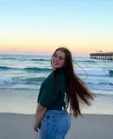 Junge Frau mit langen Haaren am Strand, dahinter Meer und eine Seebrücke im Abendlicht