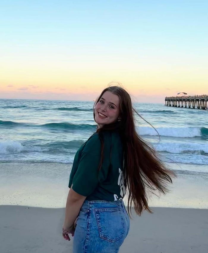 Junge Frau mit langen Haaren am Strand, dahinter Meer und eine Seebrücke im Abendlicht