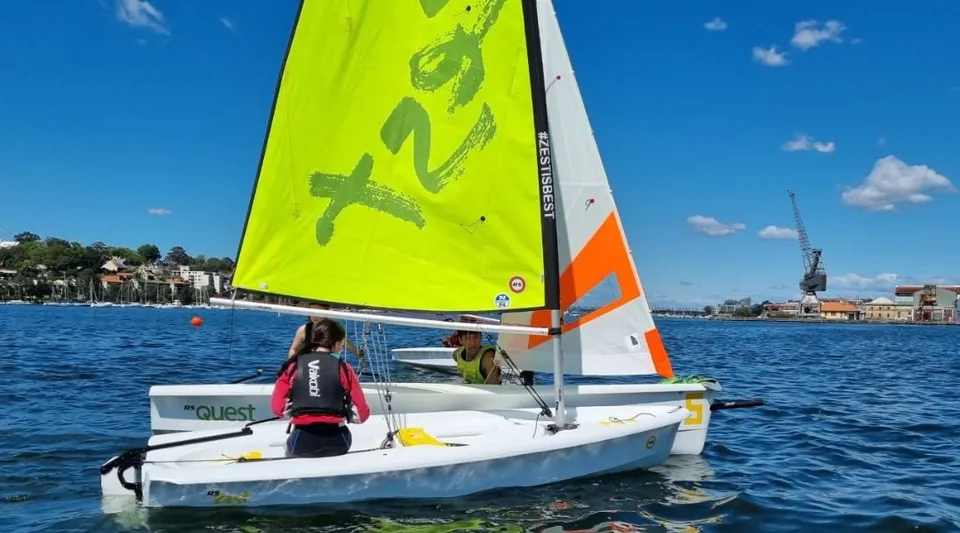 Una piccola barca a vela con vela giallo-arancione naviga sull'acqua davanti ad edifici vicino alla Hunters Hill High School.