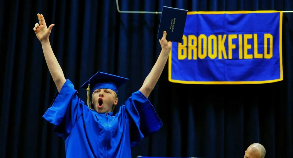 Una persona con la toga di laurea blu solleva le braccia per esultare davanti a uno striscione della Brookfield High School.