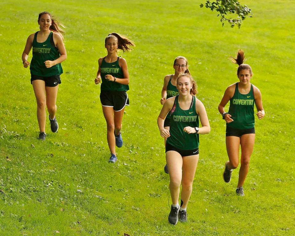 Un gruppo di giovani atlete della Coventry High School corre su un campo erboso sotto il sole.