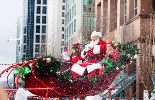 Papá Noel y dos niños disfrazados de duendes saludan sobre un trineo rojo decorado