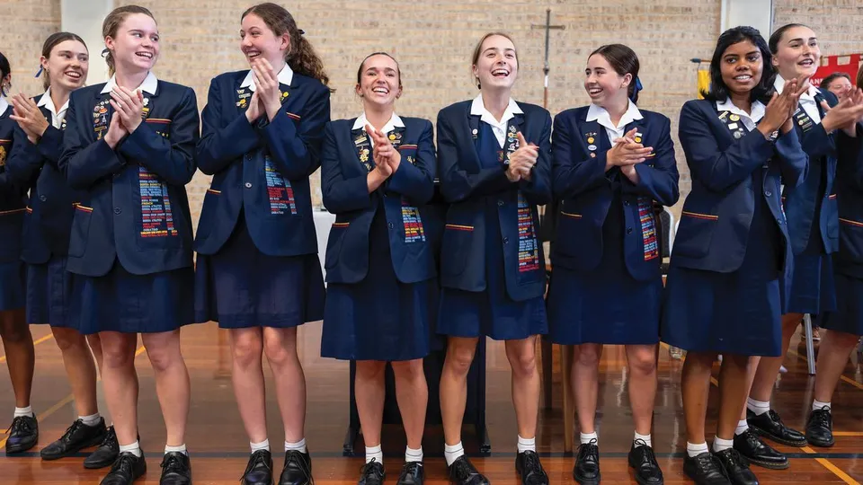 Studentesse della St. Hilda’s School in uniforme scura stanno in fila in una sala e applaudono sorridendo.