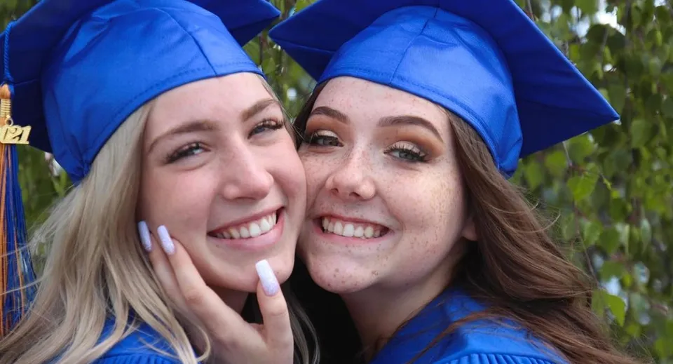 Due studentesse della Nechako Valley Secondary School in toga da diploma blu posano insieme davanti a uno sfondo naturale sfocato.