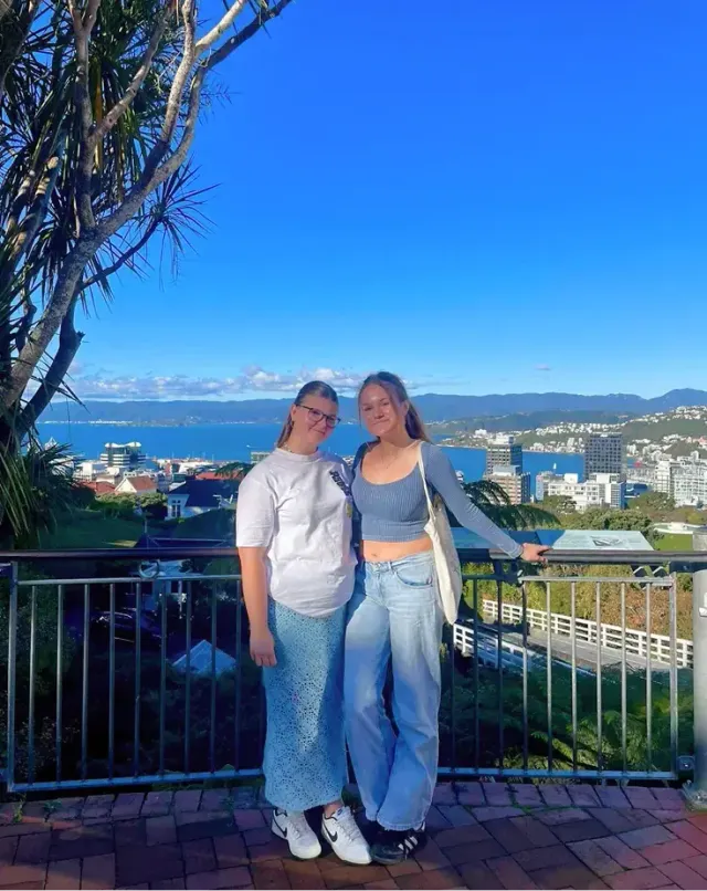 Due studentesse a un belvedere con la vista su una città portuale e la baia in Nuova Zelanda