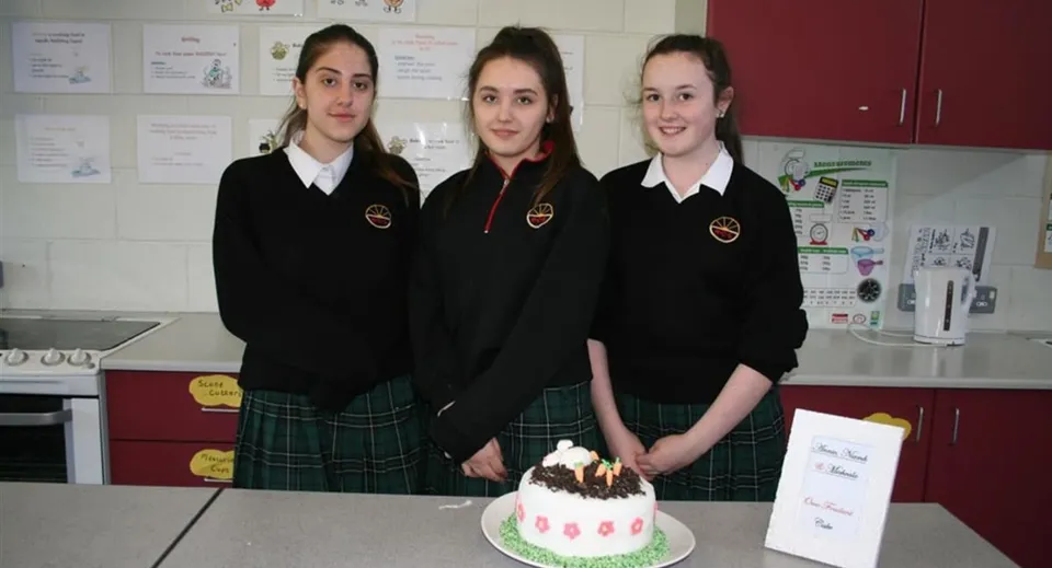 Tre studentesse della Ballinteer Community School sono insieme in classe davanti a una torta di compleanno sul tavolo.