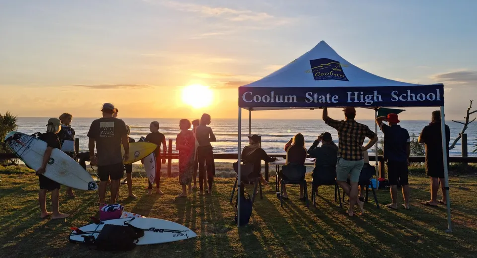Un gruppo di studenti si trova davanti allo striscione della Coolum State High School sulla spiaggia al tramonto.