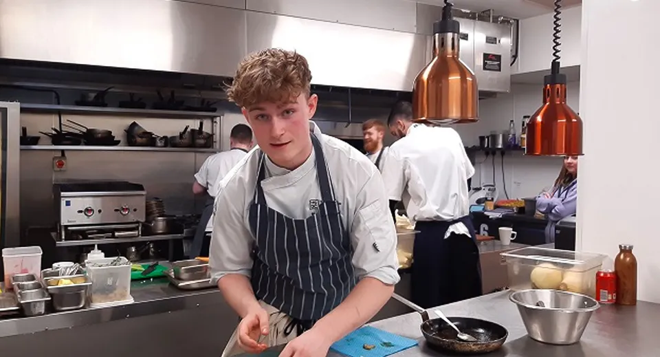 Uno studente in divisa da chef è in piedi nella cucina del campus del South Downs College circondato da utensili da cucina.