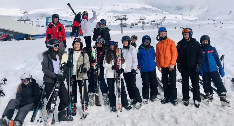 Un gruppo di studenti del Braemar College in abbigliamento invernale con attrezzatura da sci si trova su un pendio innevato davanti a montagne e impianti di risalita.