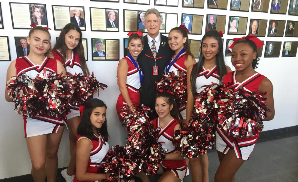 Un gruppo di cheerleader in uniformi rosse e bianche posa davanti a una parete con ritratti incorniciati alla Miami Beach Senior High School.