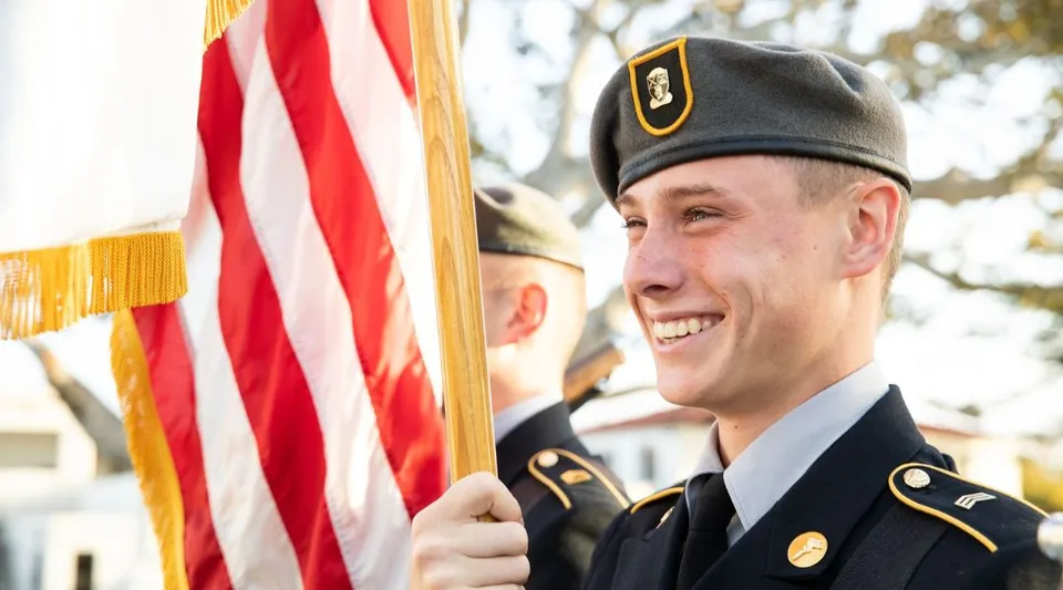 Un uomo in uniforme militare si trova accanto a una bandiera americana davanti a un paesaggio innevato nel terreno della Army and Navy Academy.