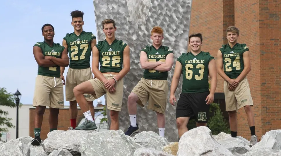 Un gruppo di atleti si trova su uno spuntone roccioso davanti a un edificio del campus della Muskegon Catholic Central High School.