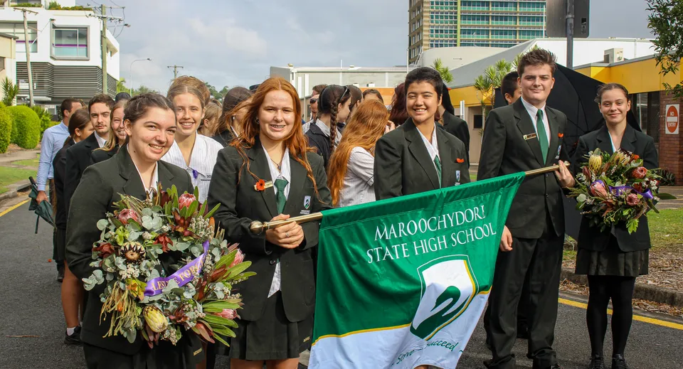 Un gruppo di studenti della Maroochydore State High School indossa la toga da diploma e tiene uno striscione davanti a dei grattacieli.