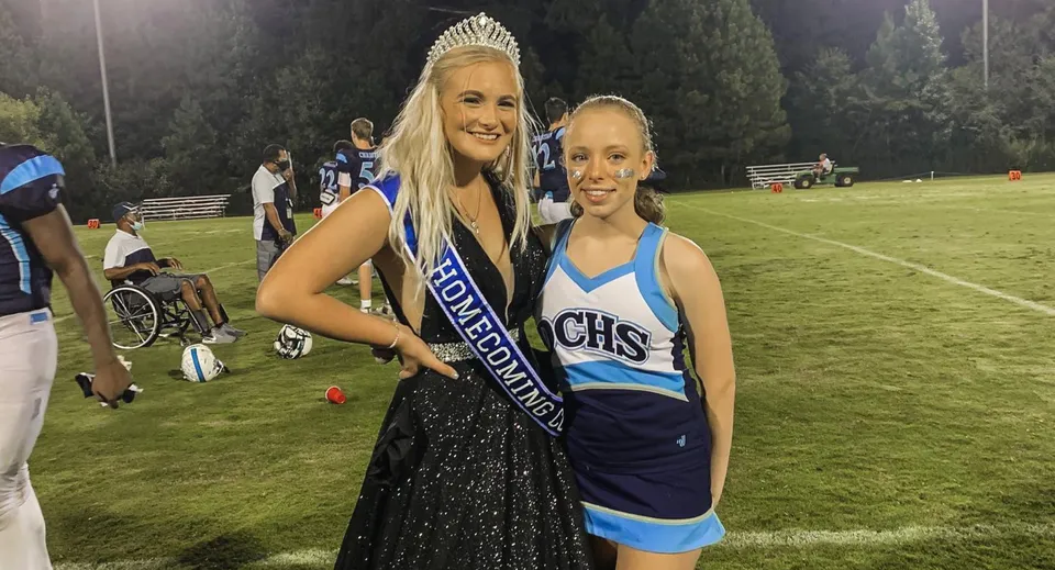 Due studentesse della Dominion Christian School si trovano di notte sul campo sportivo, una come Homecoming Queen con corona e fascia, l'altra in uniforme da cheerleader.