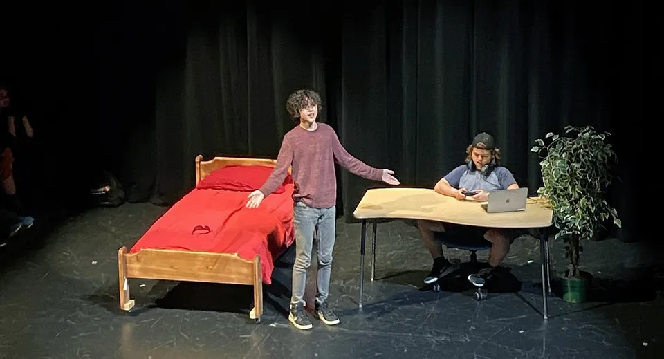 Una studentessa con una maglietta rosa sta sul palco della Imagine High Integrated Arts and Technology Secondary School con un divano rosso e una persona seduta a un tavolo sullo sfondo.