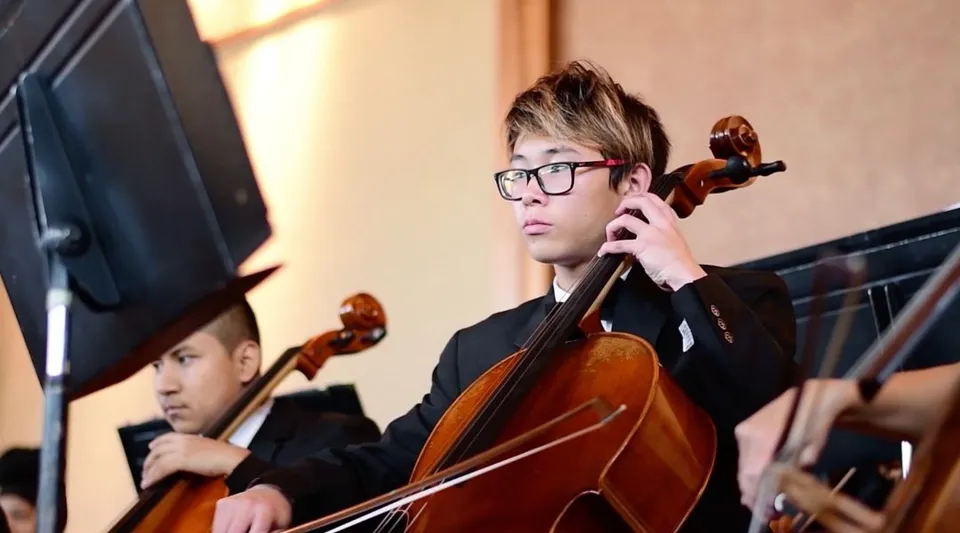 Uno studente della Auburn Adventist Academy suona il violoncello sul palco con altri musicisti sullo sfondo.