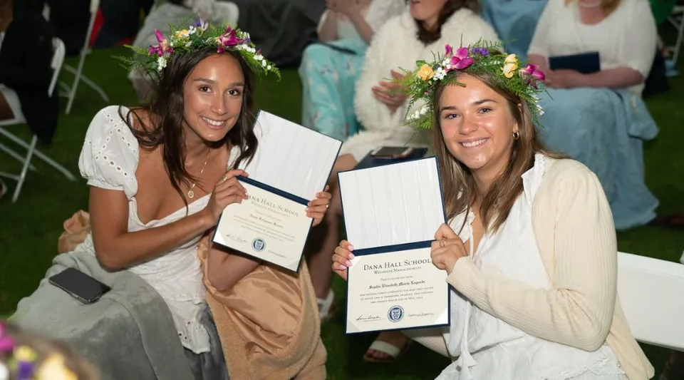 Due studentesse della Dana Hall School indossano abiti bianchi e corone di fiori e tengono in mano i loro diplomi durante una cerimonia all'aperto.