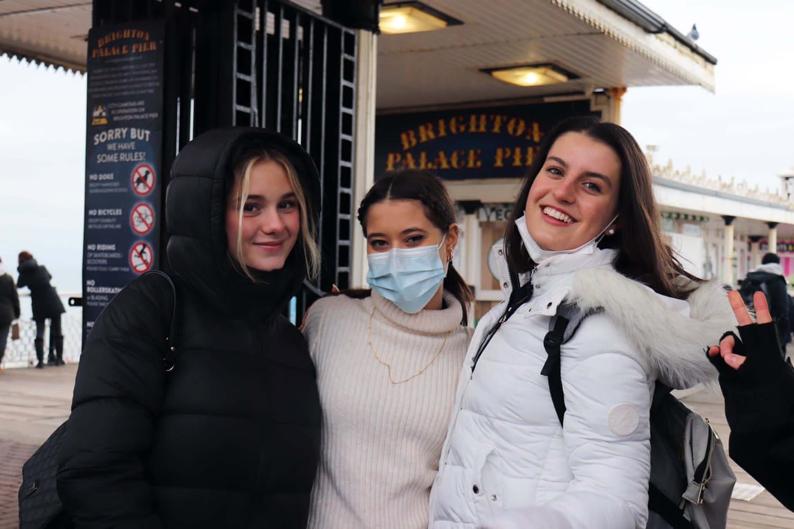 Drei junge Frauen in Winterjacken stehen lächelnd vor dem Eingang mit Schriftzug Brighton Palace Pier