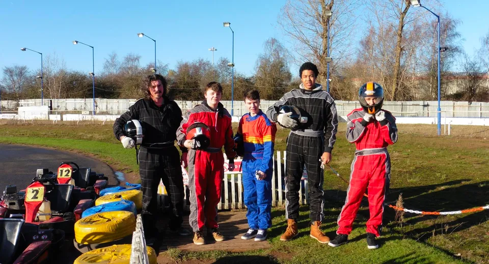 Studenti dell'Oxford Sixth Form College in tute da corsa si trovano su un circuito automobilistico davanti a un prato con alberi.