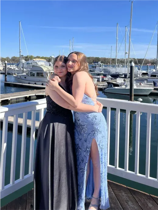 Due ragazze in abito da sera si abbracciano su un pontile davanti a un porto con barche a vela negli Stati Uniti.