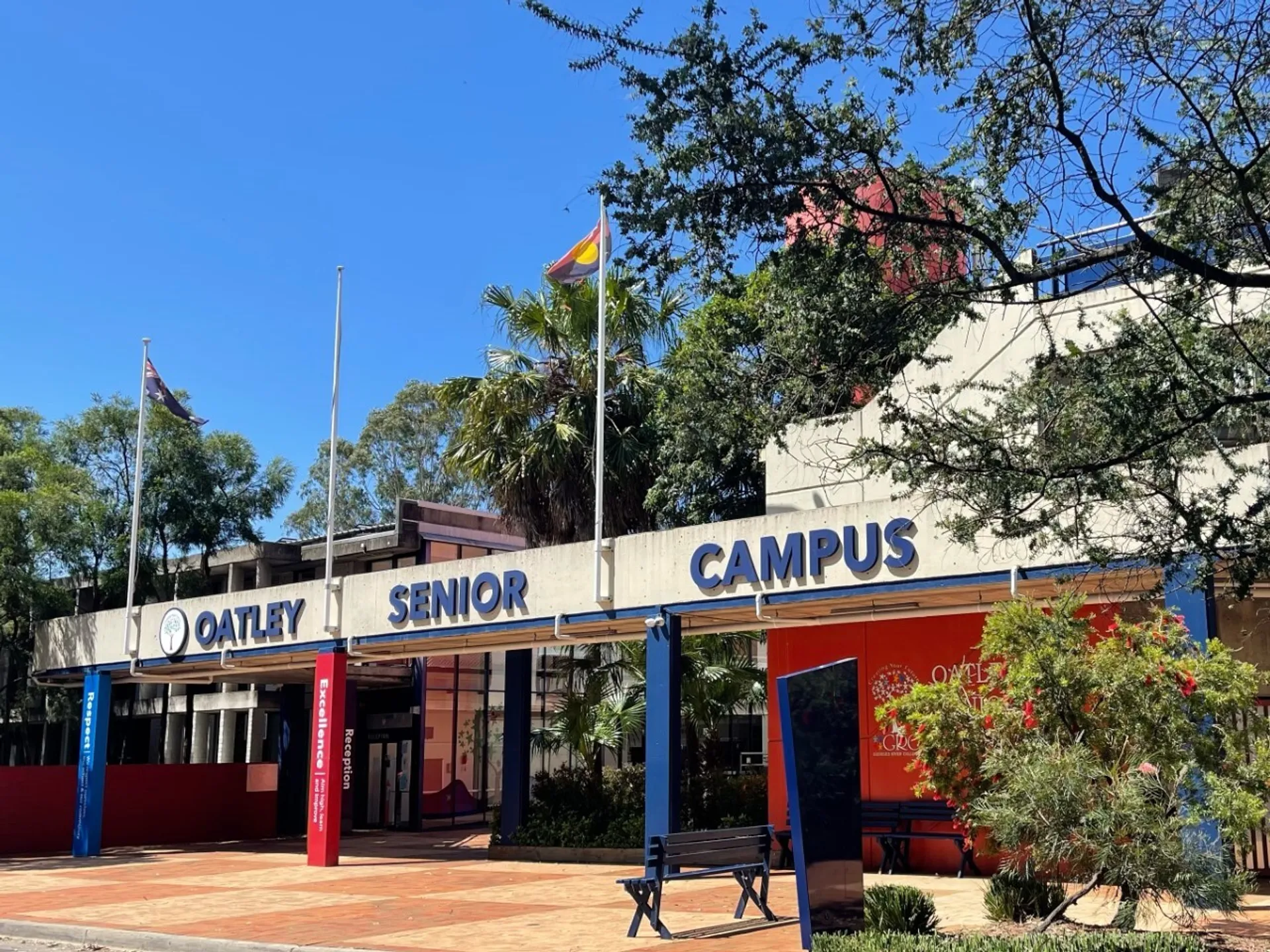 L'ingresso dell'Oatley Senior Campus del Georges River College con bandiere e panchine sotto un cielo azzurro.
