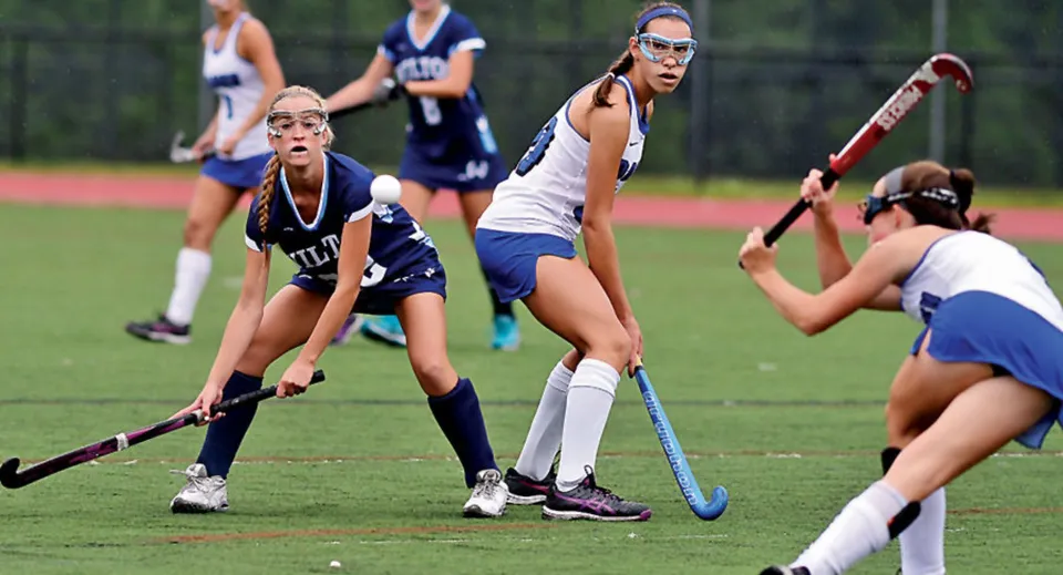 Studentesse della St. Raphaela’s Secondary School giocano una partita di hockey su un campo con erba verde e alberi sullo sfondo.