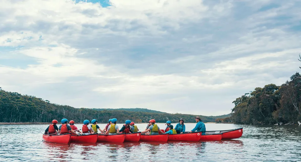 Studenti della Helensvale State High School pagaiano su una canoa rossa in un lago indossando giubbotti di salvataggio colorati, circondati da verdi colline.