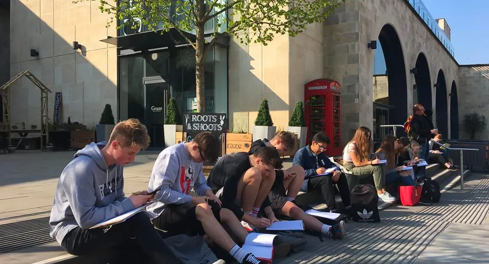 Un gruppo di giovani è seduto sul marciapiede davanti a un edificio del campus della Somerset Studio School.