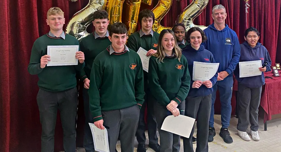 Studenti del St. Fergal’s College in divisa scolastica sono in piedi con dei certificati davanti a uno sfondo decorato in oro e lilla.