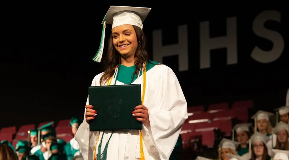 Una diplomanda sorridente della Medicine Hat High School tiene in mano il suo diploma sul palco davanti a uno sfondo con le lettere HHS.