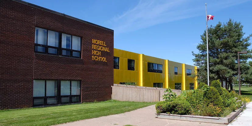 L'edificio in mattoni della Morell Regional High School con dettagli gialli sorge su un prato con alberi e una bandiera canadese che sventola in primo piano.