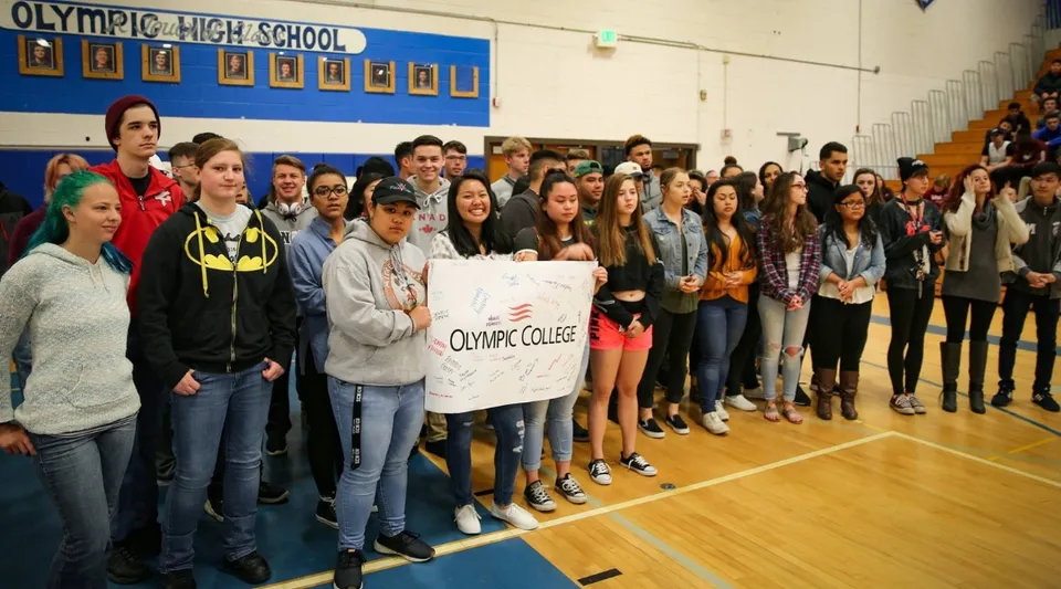 Un gruppo di studenti dell'Olympic College si riunisce in palestra e tiene un cartello con il nome della scuola.