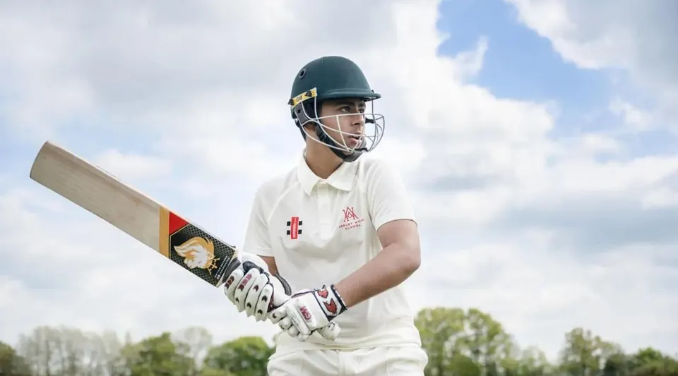 Un giocatore di cricket della Akeley Wood School si trova sul campo in divisa bianca e casco, con un cielo nuvoloso e alberi sullo sfondo.