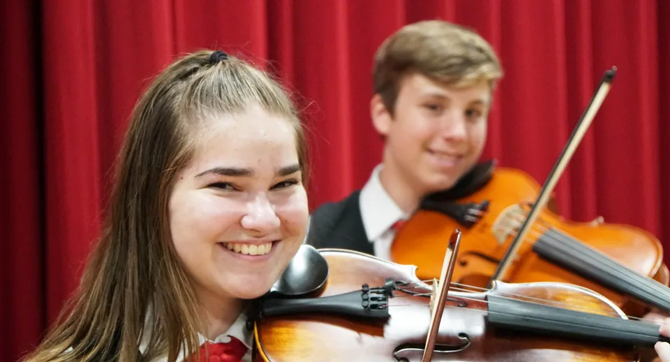 Studenti della Kirwan State High School suonano il violino insieme su un palco davanti a un sipario rosso.