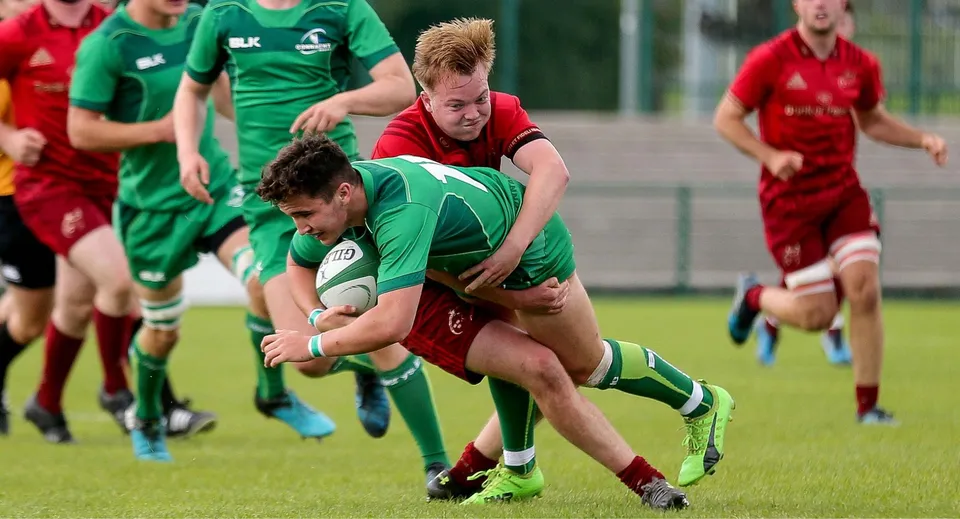 Un giocatore di rugby del Midleton College con la palla viene placcato da un avversario sul campo da gioco.