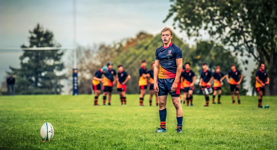 Un giocatore di rugby della Ryde School si trova su un campo d'erba davanti a un pallone da rugby con altri giocatori sullo sfondo.