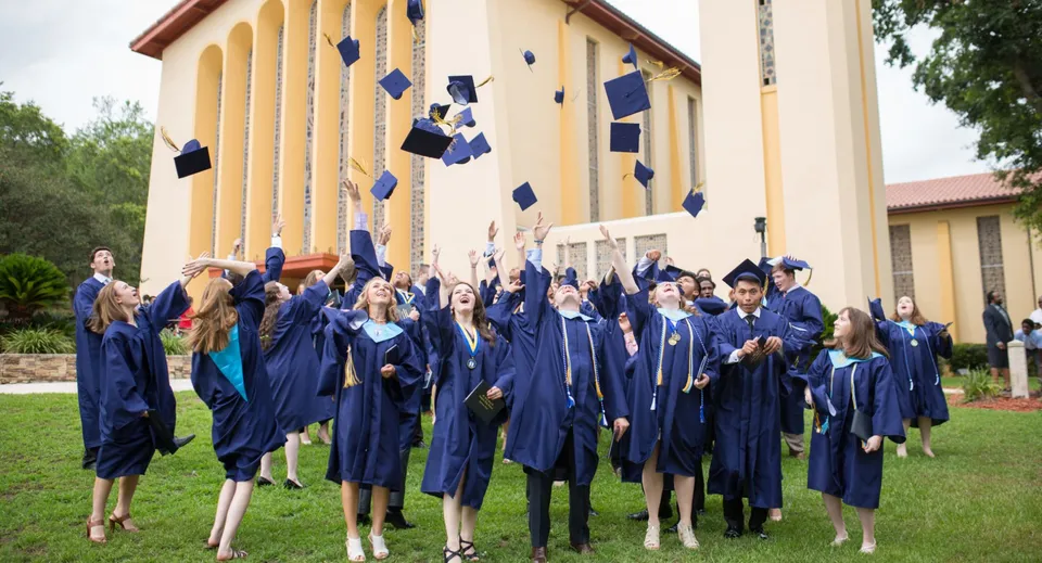 I diplomati della St. John Paul II Catholic High School lanciano in aria i loro tocchi blu davanti a un edificio giallo.