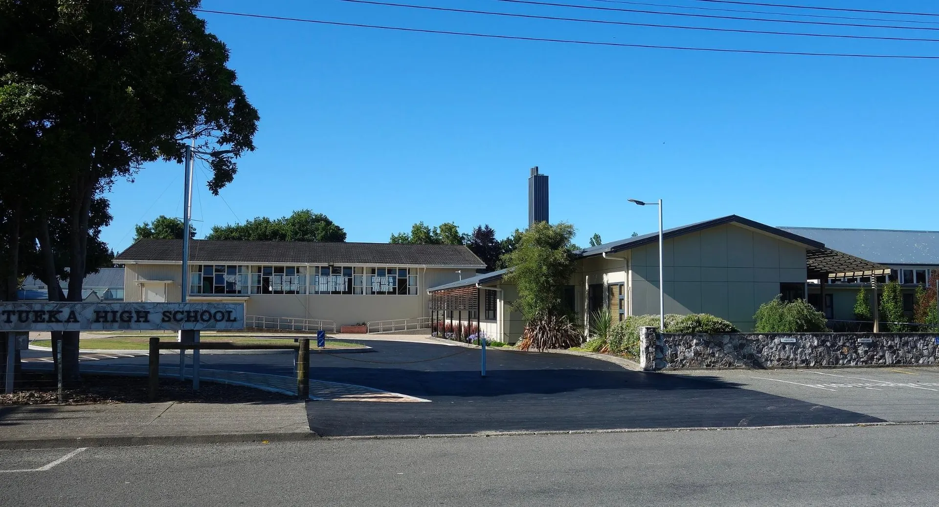 L'edificio scolastico con un'alta torre della Motueka High School si trova sullo sfondo circondato da alberi e da un parcheggio pavimentato.