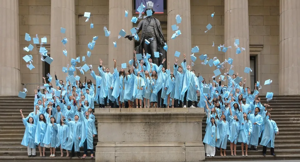 Gli studenti della Léman Manhattan Preparatory School si riuniscono con toghe blu attorno a una grande statua su una scalinata.