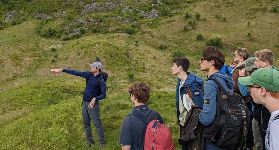 Gli studenti del Winchester College sono in piedi su una collina erbosa con un paesaggio pittoresco sullo sfondo.