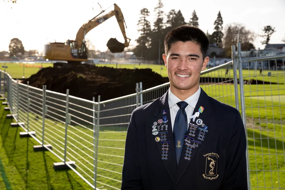 Uno studente del Tauranga Boy's College sorride in divisa davanti a un campo sportivo con macchinari edili sullo sfondo.